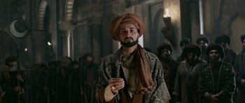 Movie still from “Padmaavat” (2018), directed by Sanjay Leela Bhansali – A man with a beard and a turban is smoking a cigarette in front of a group of people; Medium shot, Low angle