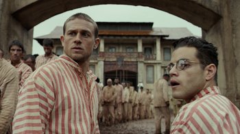 Movie still from “Papillon” (2017), directed by Michael Noer – A group of men standing next to each other in front of a building; Medium shot, Over the shoulder angle