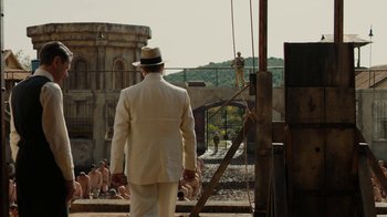 Movie still from “Papillon” (2017), directed by Michael Noer – A man in a white suit and a hat is standing in front of a fence; Wide shot, Over the shoulder angle