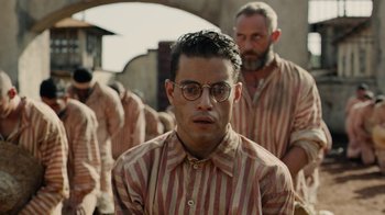 Movie still from “Papillon” (2017), directed by Michael Noer – A man in striped shirt and glasses in front of other men; Close Up shot, Over the shoulder angle