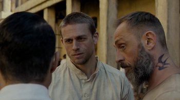 Movie still from “Papillon” (2017), directed by Michael Noer – A man in a white shirt is talking to two other men; Close Up shot, Over the shoulder angle