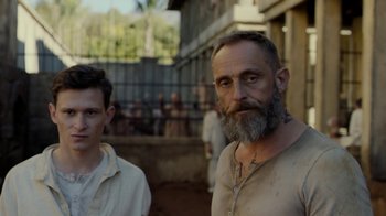Movie still from “Papillon” (2017), directed by Michael Noer – A man with a long beard standing next to another man with short hair; Close Up shot, Over the shoulder angle