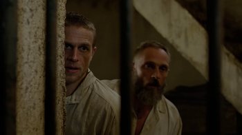 Movie still from “Papillon” (2017), directed by Michael Noer – Two men standing next to each other behind a fence; Medium shot, Over the shoulder angle