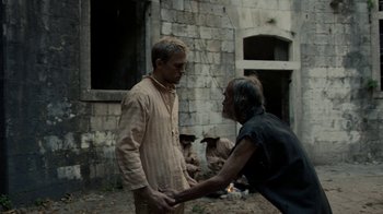 Movie still from “Papillon” (2017), directed by Michael Noer – Two men holding hands in front of an old building; Medium shot, Over the shoulder angle