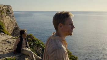 Movie still from “Papillon” (2017), directed by Michael Noer – A man standing on top of a hill next to a body of water; Medium shot, Low angle