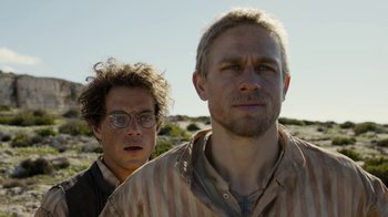 Movie still from “Papillon” (2017), directed by Michael Noer – A man standing next to another man in a field; Close Up shot, Low angle