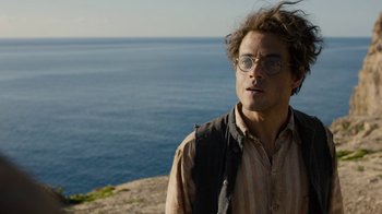 Movie still from “Papillon” (2017), directed by Michael Noer – A man standing on top of a hill near a body of water; Medium shot, Over the shoulder angle