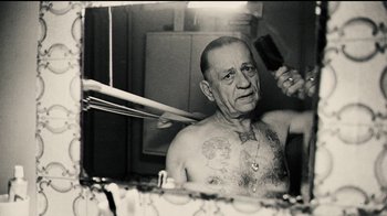 Movie still from “Papillon” (2017), directed by Michael Noer – An old man with a tattoo is combing his hair; Medium shot, Low angle