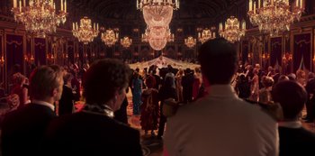 Movie still from “Paradise Hills” (2019), directed by Alice Waddington – A group of people standing in front of chandeliers in a room; Extreme Wide shot, Over the shoulder angle