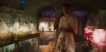 Movie still from “Paradise Hills” (2019), directed by Alice Waddington – A man standing in front of a table with plants on it; Medium shot, Low angle