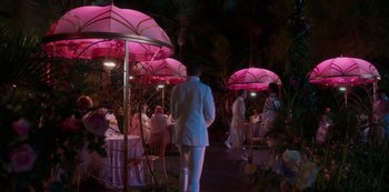 Movie still from “Paradise Hills” (2019), directed by Alice Waddington – A group of people standing around tables with pink umbrellas over them; Extreme Wide shot, High angle