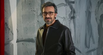 Movie still from “Parallel Mothers” (2021), directed by Pedro Almodóvar – A man wearing a black leather jacket and glasses standing in front of a gray wall; Medium shot, Low angle