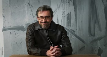 Movie still from “Parallel Mothers” (2021), directed by Pedro Almodóvar – A man wearing glasses and a leather jacket sitting at a table with his hands crossed; Medium shot, Low angle