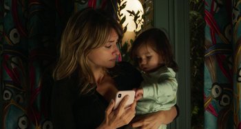 Movie still from “Parallel Mothers” (2021), directed by Pedro Almodóvar – A woman holding a child while looking at a cell phone; Close Up shot, High angle