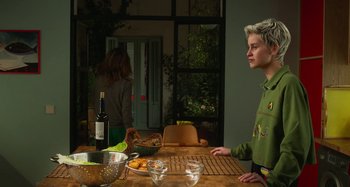 Movie still from “Parallel Mothers” (2021), directed by Pedro Almodóvar – A man standing in front of a wooden table with a bottle of wine on top of it; Medium shot, Over the shoulder angle
