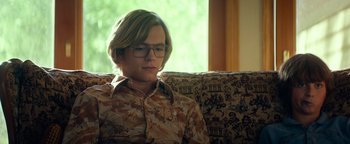 Movie still from “My Friend Dahmer” (2017), directed by Marc Meyers – A man sitting on top of a couch wearing glasses; Medium shot, Over the shoulder angle