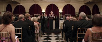 Movie still from “Parker” (2013), directed by Taylor Hackford – A man standing at a podium in front of a group of people in tuxedos; Wide shot, Over the shoulder angle