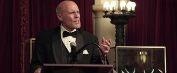 Movie still from “Parker” (2013), directed by Taylor Hackford – An older man in a tuxedo speaking into a microphone; Medium shot, Low angle