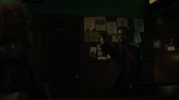 Movie still from “Paterson” (2016), directed by Jim Jarmusch – A man holding a handgun while standing in front of a bulletin board; Medium shot, Over the shoulder angle