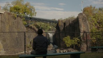 Movie still from “Paterson” (2016), directed by Jim Jarmusch – A man sitting on a bench in front of a waterfall; Extreme Wide shot, High angle