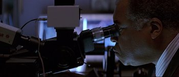 Movie still from “Patriot Games” (1992), directed by Phillip Noyce – A man looking through a microscope at something on a table; Close Up shot, Low angle