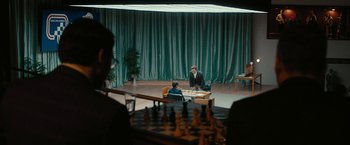 Movie still from “Pawn Sacrifice” (2014), directed by Edward Zwick – Two people playing chess on a stage with green drapes; Extreme Wide shot, Over the shoulder angle