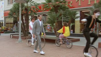Movie still from “Pee-wee's Big Adventure” (1985), directed by Tim Burton – A man in a suit is walking down the street while a woman is riding a bike; Wide shot, Low angle