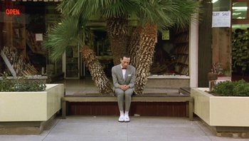Movie still from “Pee-wee's Big Adventure” (1985), directed by Tim Burton – A man sitting on a bench in front of some palm trees; Wide shot, High angle