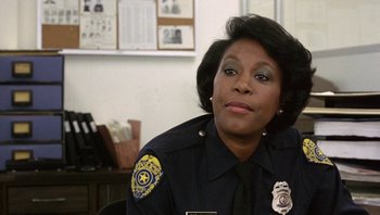 Movie still from “Pee-wee's Big Adventure” (1985), directed by Tim Burton – A police officer sitting at a desk in front of a picture; Close Up shot, Over the shoulder angle