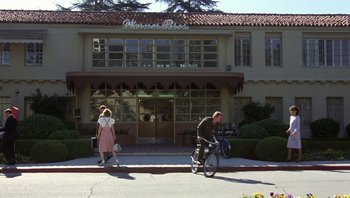 Movie still from “Pee-wee's Big Adventure” (1985), directed by Tim Burton – A man riding a bike down a street next to a building; Extreme Wide shot, Low angle