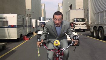 Movie still from “Pee-wee's Big Adventure” (1985), directed by Tim Burton – A man riding a bike down a street with other vehicles behind him; Wide shot, Over the shoulder angle