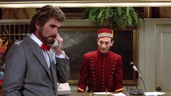 Movie still from “Pee-wee's Big Adventure” (1985), directed by Tim Burton – A man talking on a phone while a man in a red suit stands in front of a microphone; Medium shot, Over the shoulder angle