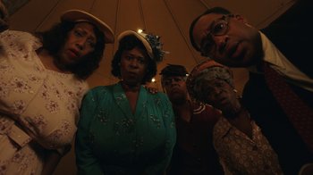 Movie still from “A League of Their Own” (2022), created by Abbi Jacobson – A group of people that are standing under an umbrella; Medium shot, Low angle