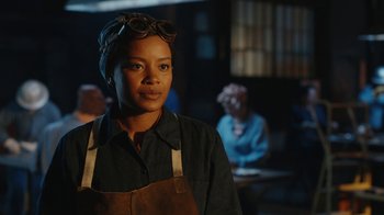 Movie still from “A League of Their Own” (2022), created by Abbi Jacobson – A woman wearing an overalls and glasses in front of a group of people; Medium shot, Over the shoulder angle