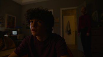 Movie still from “Full Circle” (2023), directed by Steven Soderbergh – A young man sitting at a table in a room; Medium shot, Over the shoulder angle