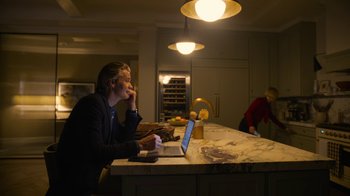 Movie still from “Full Circle” (2023), directed by Steven Soderbergh – A man sitting in front of a laptop computer in a kitchen; Medium shot, Over the shoulder angle
