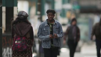 Movie still from “Full Circle” (2023), directed by Steven Soderbergh – A young man is standing on the sidewalk looking at his cell phone; Medium shot, Over the shoulder angle