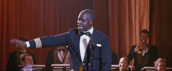 Movie still from “Poseidon” (2006), directed by Wolfgang Petersen – A man wearing a suit and bow tie is holding a microphone; Medium shot, Low angle