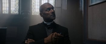 Movie still from “Prey for the Devil” (2022), directed by Daniel Stamm – A man in a priest's outfit smoking a cigarette; Medium shot, Low angle