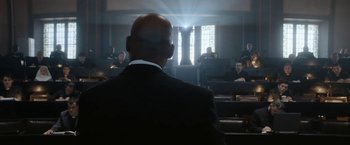 Movie still from “Prey for the Devil” (2022), directed by Daniel Stamm – A man in a black suit is sitting in front of a group of people; Wide shot, Low angle