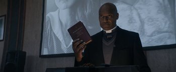 Movie still from “Prey for the Devil” (2022), directed by Daniel Stamm – A man in a priest's outfit holding a bible; Close Up shot, Low angle