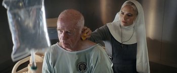 Movie still from “Prey for the Devil” (2022), directed by Daniel Stamm – An older man getting his ears brushed by a nun; Medium shot, Over the shoulder angle