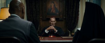 Movie still from “Prey for the Devil” (2022), directed by Daniel Stamm – A man sitting at a table in front of a painting; Medium shot, Over the shoulder angle