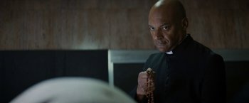 Movie still from “Prey for the Devil” (2022), directed by Daniel Stamm – A man in a priest's outfit holding a rosary; Close Up shot, Low angle