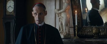 Movie still from “Prey for the Devil” (2022), directed by Daniel Stamm – A man in a red and black outfit standing in front of a statue; Close Up shot, Over the shoulder angle