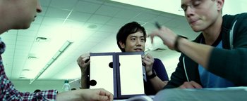 Movie still from “Project Almanac” (2015), directed by Dean Israelite – A man holding a binder in front of a group of people; Medium shot, Over the shoulder angle