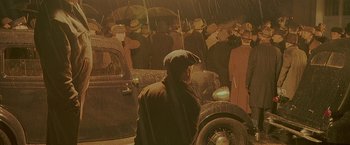 Movie still from “Public Enemies” (2009), directed by Michael Mann – A man in a hat and a coat standing next to a car in the rain with an umbrella; Wide shot, High angle