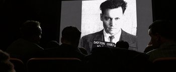 Movie still from “Public Enemies” (2009), directed by Michael Mann – A man in a suit and tie sitting in front of a picture of johnny depp; Close Up shot, Over the shoulder angle