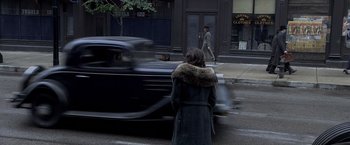 Movie still from “Public Enemies” (2009), directed by Michael Mann – A woman in a fur lined coat is walking down the street past an old car; Wide shot, Over the shoulder angle