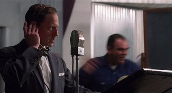 Movie still from “Quiz Show” (1994), directed by Robert Redford – A man standing in front of a microphone in front of another man; Medium shot, Low angle